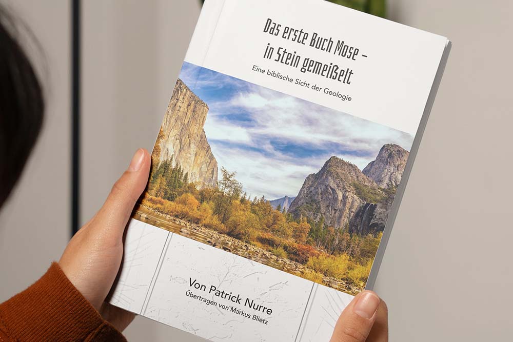 Abbildung des Buches "Das erste Buch Mose in Stein gemeißelt" in der Hand einer Frau. Die Vorderseite des Bildes zeigt Berge, grün-gelber Wald und einen Fluss.